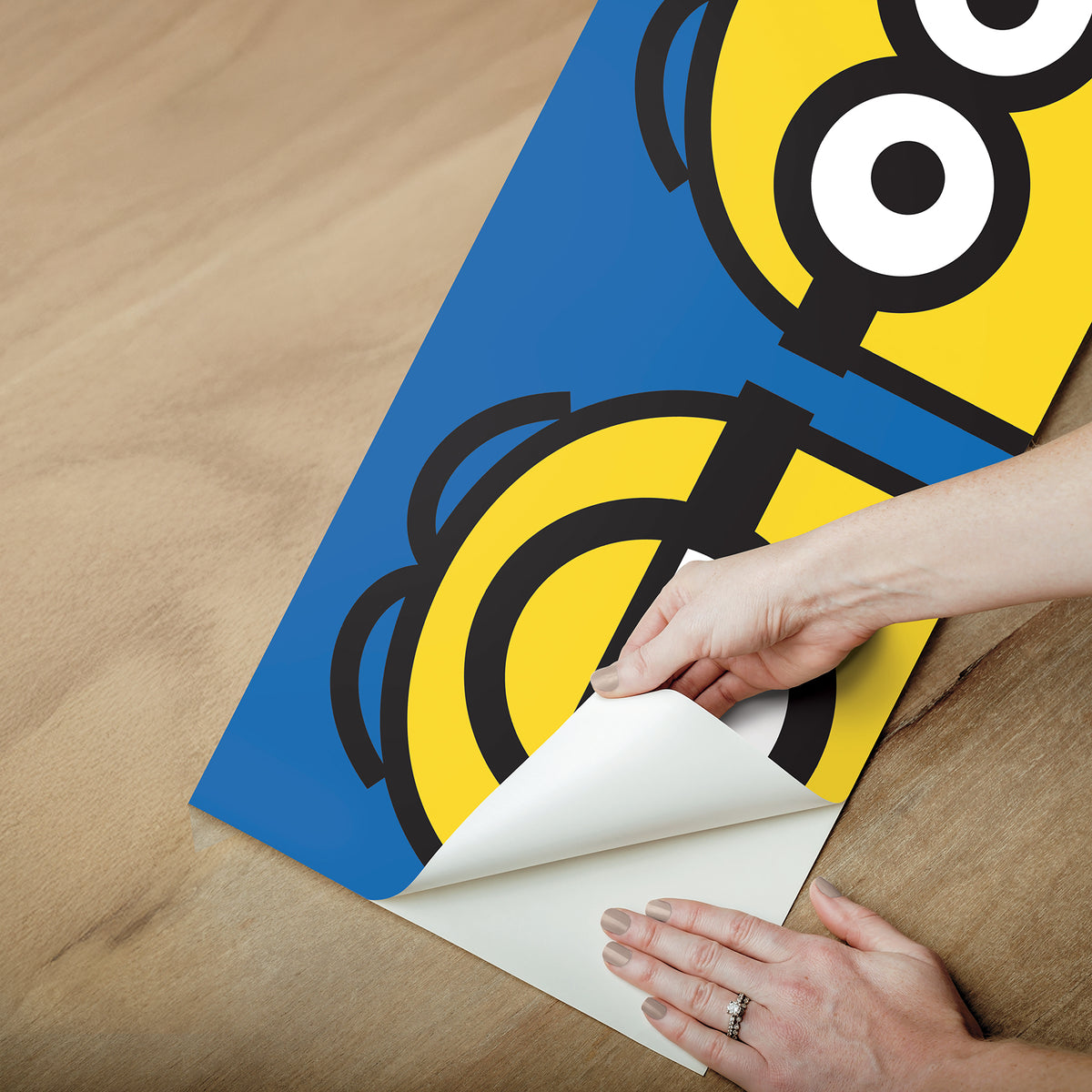 A person wearing a silver ring on their hand peels the backing off a large sticker resembling the WallPops Universal Studios Minions Always Eyeconic Border. The sticker features part of a cartoon character with yellow skin, round eyes, and black goggles set against a blue and yellow background, all laid out on a wooden surface.