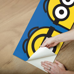 A person wearing a silver ring on their hand peels the backing off a large sticker resembling the WallPops Universal Studios Minions Always Eyeconic Border. The sticker features part of a cartoon character with yellow skin, round eyes, and black goggles set against a blue and yellow background, all laid out on a wooden surface.