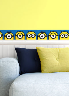 A person wearing a silver ring on their hand peels the backing off a large sticker resembling the WallPops Universal Studios Minions Always Eyeconic Border. The sticker features part of a cartoon character with yellow skin, round eyes, and black goggles set against a blue and yellow background, all laid out on a wooden surface.