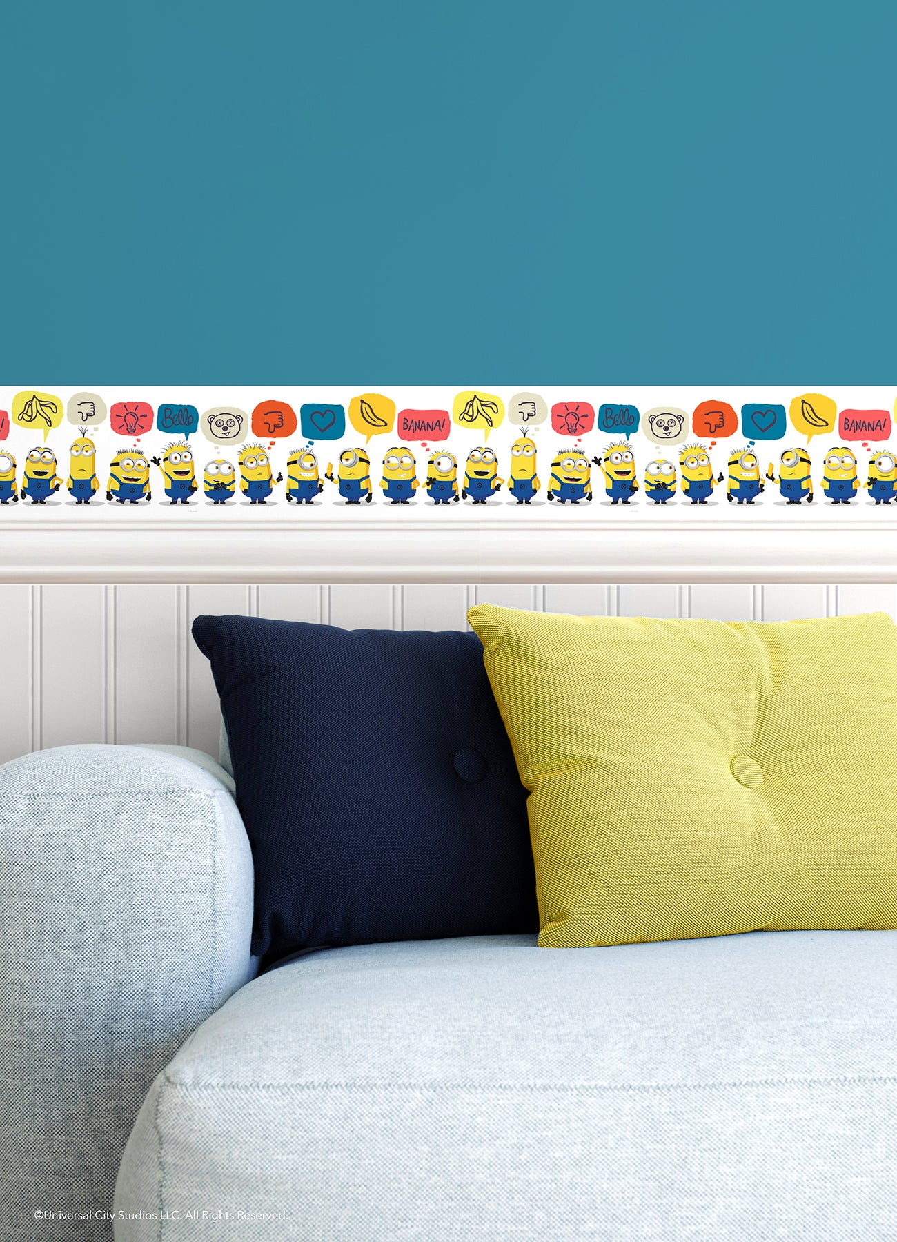 A person peels back a sheet from the WallPops Universal Studios Minions Better Together Border on a rustic wooden surface. The border features bright, playful designs including Minions characters, cartoon bananas, hearts, thumbs down icons, the word “BANANA,” and bear faces.