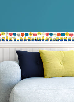 A person peels back a sheet from the WallPops Universal Studios Minions Better Together Border on a rustic wooden surface. The border features bright, playful designs including Minions characters, cartoon bananas, hearts, thumbs down icons, the word “BANANA,” and bear faces.