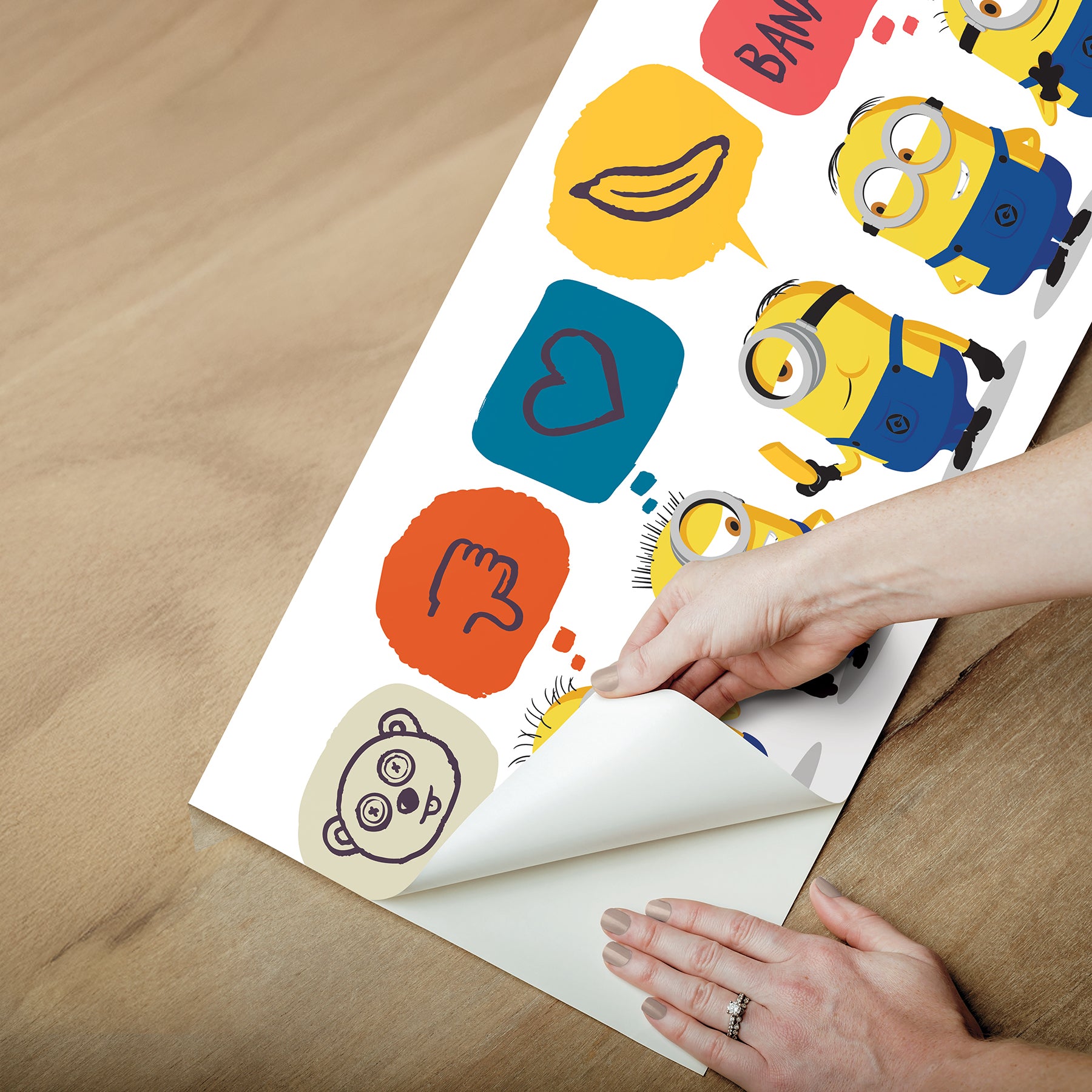 A person peels back a sheet from the WallPops Universal Studios Minions Better Together Border on a rustic wooden surface. The border features bright, playful designs including Minions characters, cartoon bananas, hearts, thumbs down icons, the word “BANANA,” and bear faces.