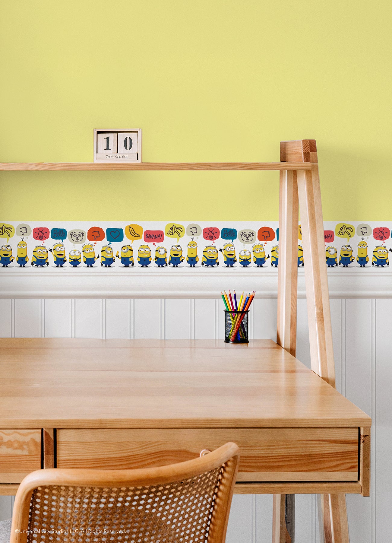 A person peels back a sheet from the WallPops Universal Studios Minions Better Together Border on a rustic wooden surface. The border features bright, playful designs including Minions characters, cartoon bananas, hearts, thumbs down icons, the word “BANANA,” and bear faces.