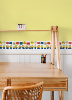 A person peels back a sheet from the WallPops Universal Studios Minions Better Together Border on a rustic wooden surface. The border features bright, playful designs including Minions characters, cartoon bananas, hearts, thumbs down icons, the word “BANANA,” and bear faces.