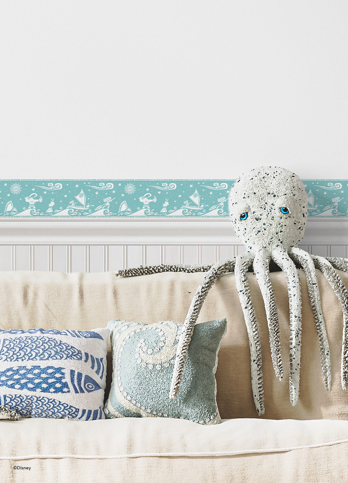 A bathroom with white cabinets, a round sink, and a wood-framed mirror. A plant in a basket, folded towels, and seashells are beside the sink. The blue wall features the Disney Moana Sail By The Stars Border by WallPops, adding whimsical aqua and white ocean motifs with playful peel-and-stick decals. Light streams in from the right.