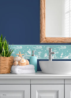 A bathroom with white cabinets, a round sink, and a wood-framed mirror. A plant in a basket, folded towels, and seashells are beside the sink. The blue wall features the Disney Moana Sail By The Stars Border by WallPops, adding whimsical aqua and white ocean motifs with playful peel-and-stick decals. Light streams in from the right.