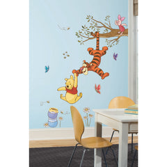 Winnie the Pooh dangles from Tigger’s tail, reaching for a falling honey pot, while Tigger hangs upside down from a tree branch as Piglet looks on above. Blue and red butterflies flutter nearby, and daisies grow below, creating a cheerful scene. The Disney Winnie the Pooh Swinging for Honey Giant Wall Decals by WallPops features these charming characters with rustic design elements—perfect for peel and stick wall decals or as accents on distressed barn wood décor to bring warmth to any room.