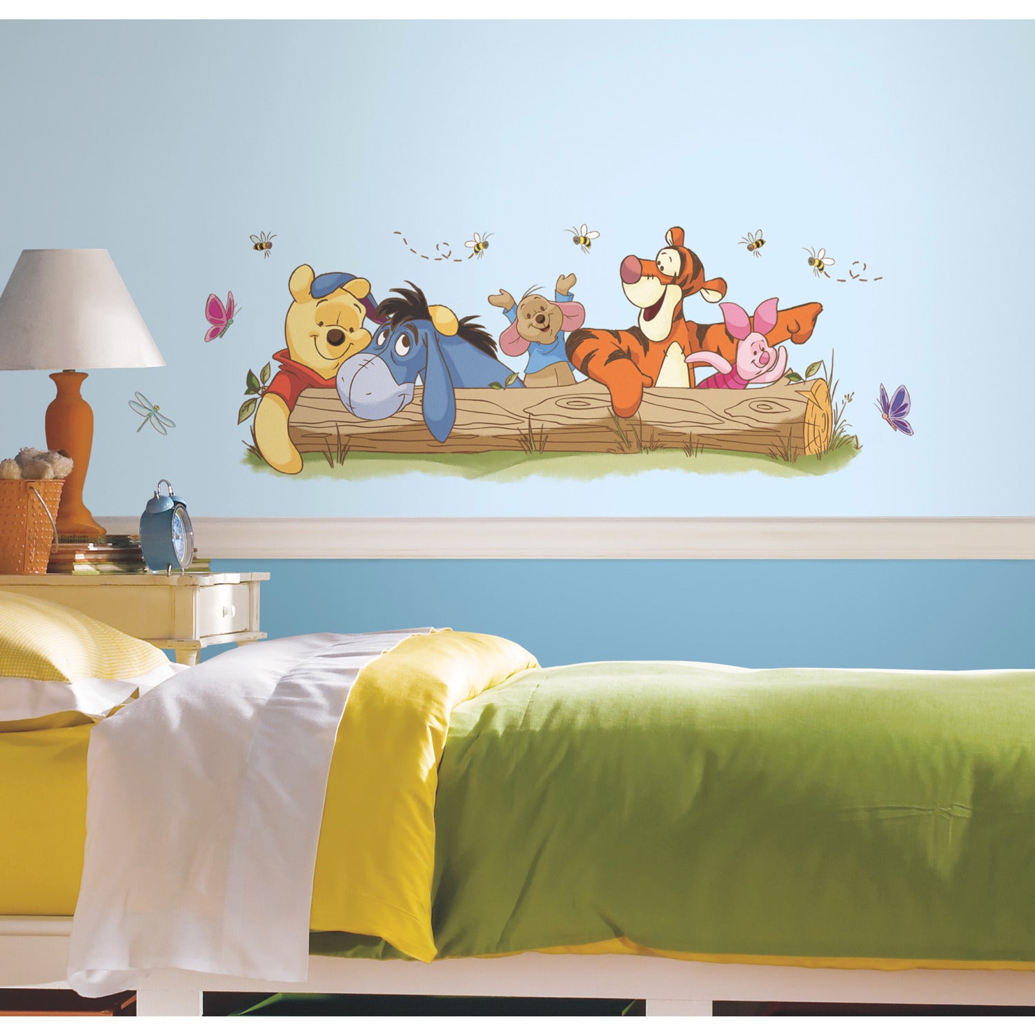 A child’s bedroom with a bed featuring yellow and green bedding. On the light blue wall, Disney Winnie the Pooh Pooh and Friends Outdoor Fun Giant Wall Decals by WallPops display large peel-and-stick images of Winnie the Pooh, Eeyore, Piglet, Tigger, and Roo leaning on a log surrounded by butterflies and bees. A white lamp and clock are placed on the bedside table.