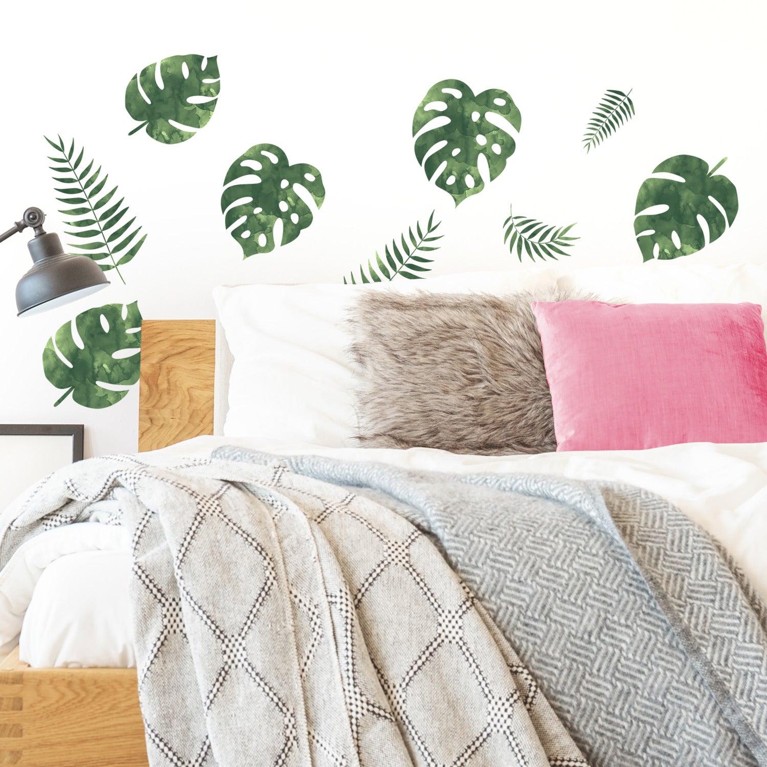 A bed featuring white and gray blankets, a pink pillow, and a furry gray pillow creates a cozy retreat. Above the bed, WallPops Palm Leaves Wall Decals add vibrant green accents to the white wall, while the rustic look of barn wood wall decals can beautifully complement the space. A black wall lamp is mounted on the left side above the wooden headboard, providing a touch of modern contrast.