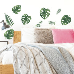 A bed featuring white and gray blankets, a pink pillow, and a furry gray pillow creates a cozy retreat. Above the bed, WallPops Palm Leaves Wall Decals add vibrant green accents to the white wall, while the rustic look of barn wood wall decals can beautifully complement the space. A black wall lamp is mounted on the left side above the wooden headboard, providing a touch of modern contrast.