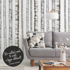 A modern bedroom features a wooden bed with crisp white bedding, a plush fur throw, potted plants, a wooden side table, and open shelving displaying both décor and additional greenery. The pale walls are adorned with the Realistic Birch Trees Peel and Stick Giant Wall Decals by WallPops, creating a serene birch tree mural that enhances the room’s natural ambiance. Subtle accents of distressed barn wood infuse rustic charm into this bright and minimal space.
