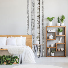 A modern bedroom features a wooden bed with crisp white bedding, a plush fur throw, potted plants, a wooden side table, and open shelving displaying both décor and additional greenery. The pale walls are adorned with the Realistic Birch Trees Peel and Stick Giant Wall Decals by WallPops, creating a serene birch tree mural that enhances the room’s natural ambiance. Subtle accents of distressed barn wood infuse rustic charm into this bright and minimal space.