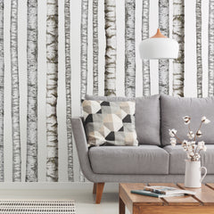 A modern bedroom features a wooden bed with crisp white bedding, a plush fur throw, potted plants, a wooden side table, and open shelving displaying both décor and additional greenery. The pale walls are adorned with the Realistic Birch Trees Peel and Stick Giant Wall Decals by WallPops, creating a serene birch tree mural that enhances the room’s natural ambiance. Subtle accents of distressed barn wood infuse rustic charm into this bright and minimal space.