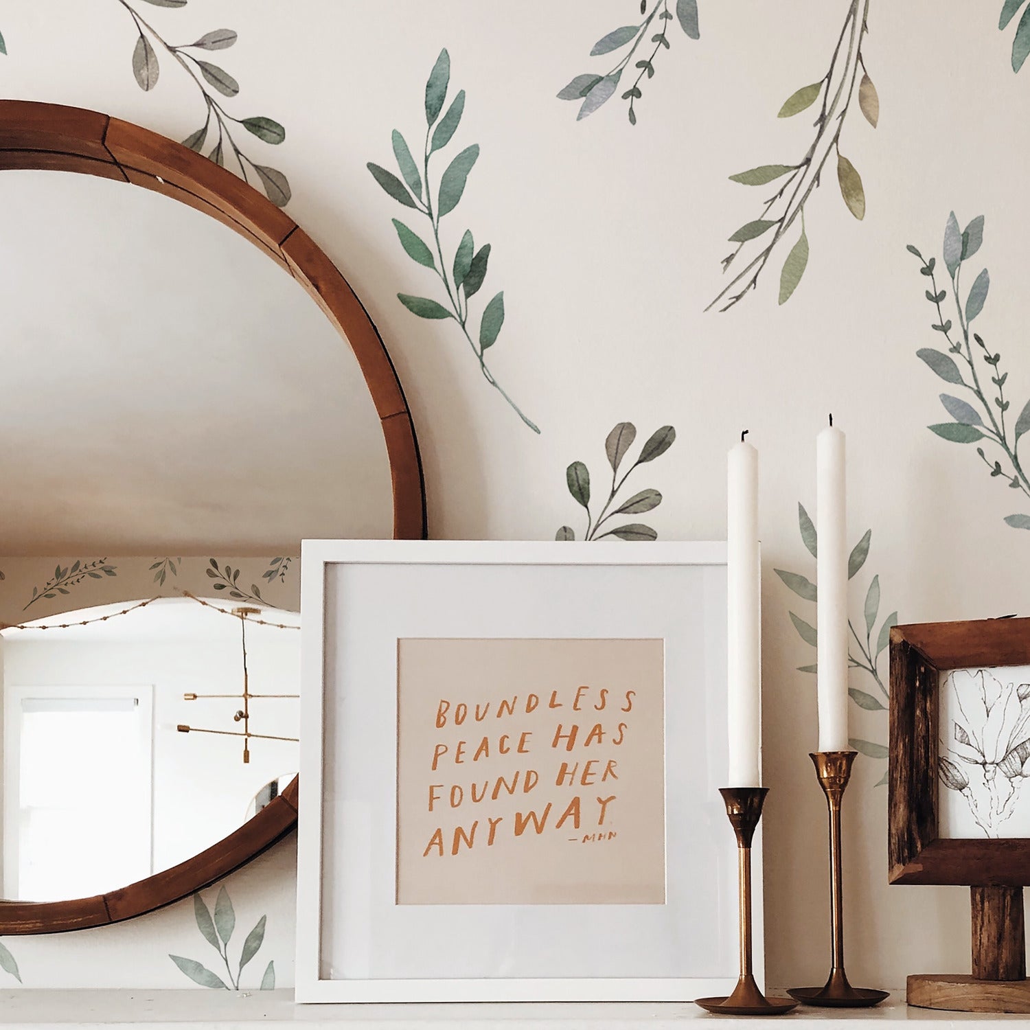 A round wooden mirror, framed quote art reading BOUNDLESS PEACE HAS FOUND HER ANYWAY, two white candles in brass holders, and a small framed drawing sit on a white shelf against a wall adorned with Country Leaves Peel and Stick Wall Decals by WallPops. The rustic design is complemented by distressed barn wood accents.