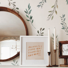 A round wooden mirror, framed quote art reading BOUNDLESS PEACE HAS FOUND HER ANYWAY, two white candles in brass holders, and a small framed drawing sit on a white shelf against a wall adorned with Country Leaves Peel and Stick Wall Decals by WallPops. The rustic design is complemented by distressed barn wood accents.