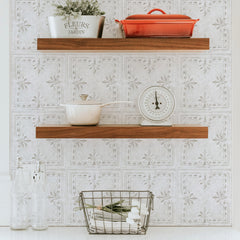 A wooden tray with rustic design rests on a white bed, holding a cup of black coffee on a saucer, a small white creamer, and a white vase with dried grass. The tray features a decorative base reminiscent of the WallPops White Tin Tile Backsplash Peel and Stick Giant Wall Decals, showcasing embossed patterns that evoke the charm of distressed barn wood.