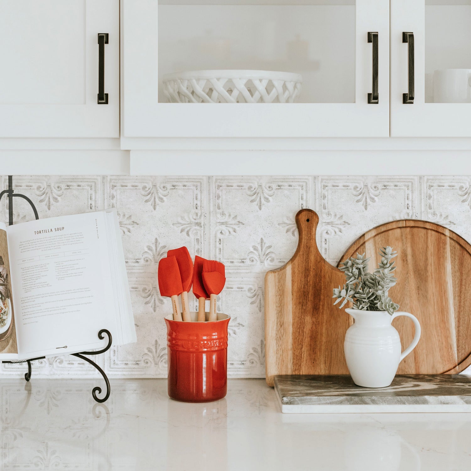 A wooden tray with rustic design rests on a white bed, holding a cup of black coffee on a saucer, a small white creamer, and a white vase with dried grass. The tray features a decorative base reminiscent of the WallPops White Tin Tile Backsplash Peel and Stick Giant Wall Decals, showcasing embossed patterns that evoke the charm of distressed barn wood.