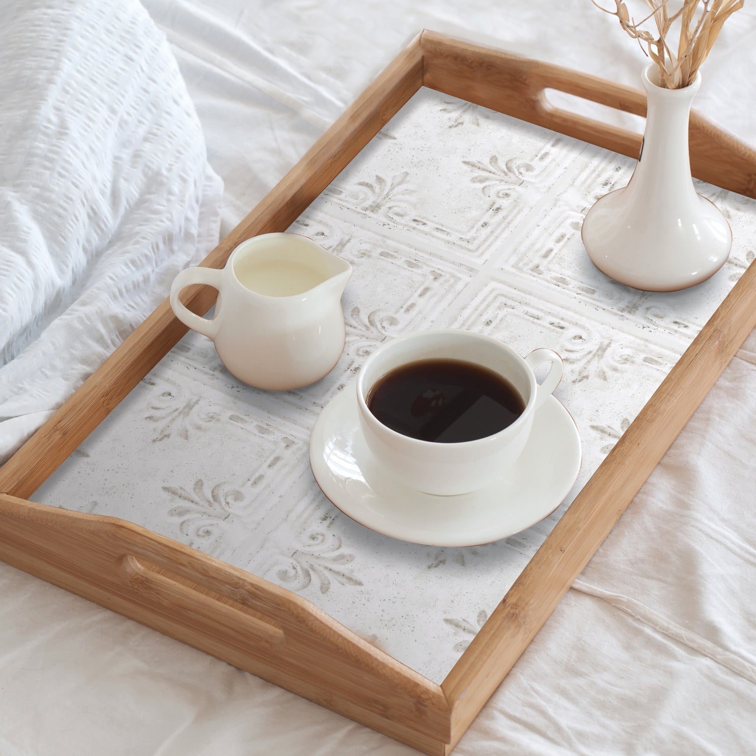 A wooden tray with rustic design rests on a white bed, holding a cup of black coffee on a saucer, a small white creamer, and a white vase with dried grass. The tray features a decorative base reminiscent of the WallPops White Tin Tile Backsplash Peel and Stick Giant Wall Decals, showcasing embossed patterns that evoke the charm of distressed barn wood.