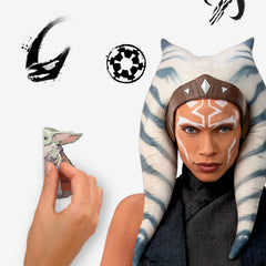 A striking silhouette of Ahsoka Tano, clad in black armor with blue and white striped head-tails and facial markings, stands confidently holding two crossed white lightsabers—her arms wrapped in dark gauntlets—against a plain white background. The Star Wars Ahsoka Peel and Stick Giant Wall Decal by WallPops is an inspiring way to add bold character to any room.