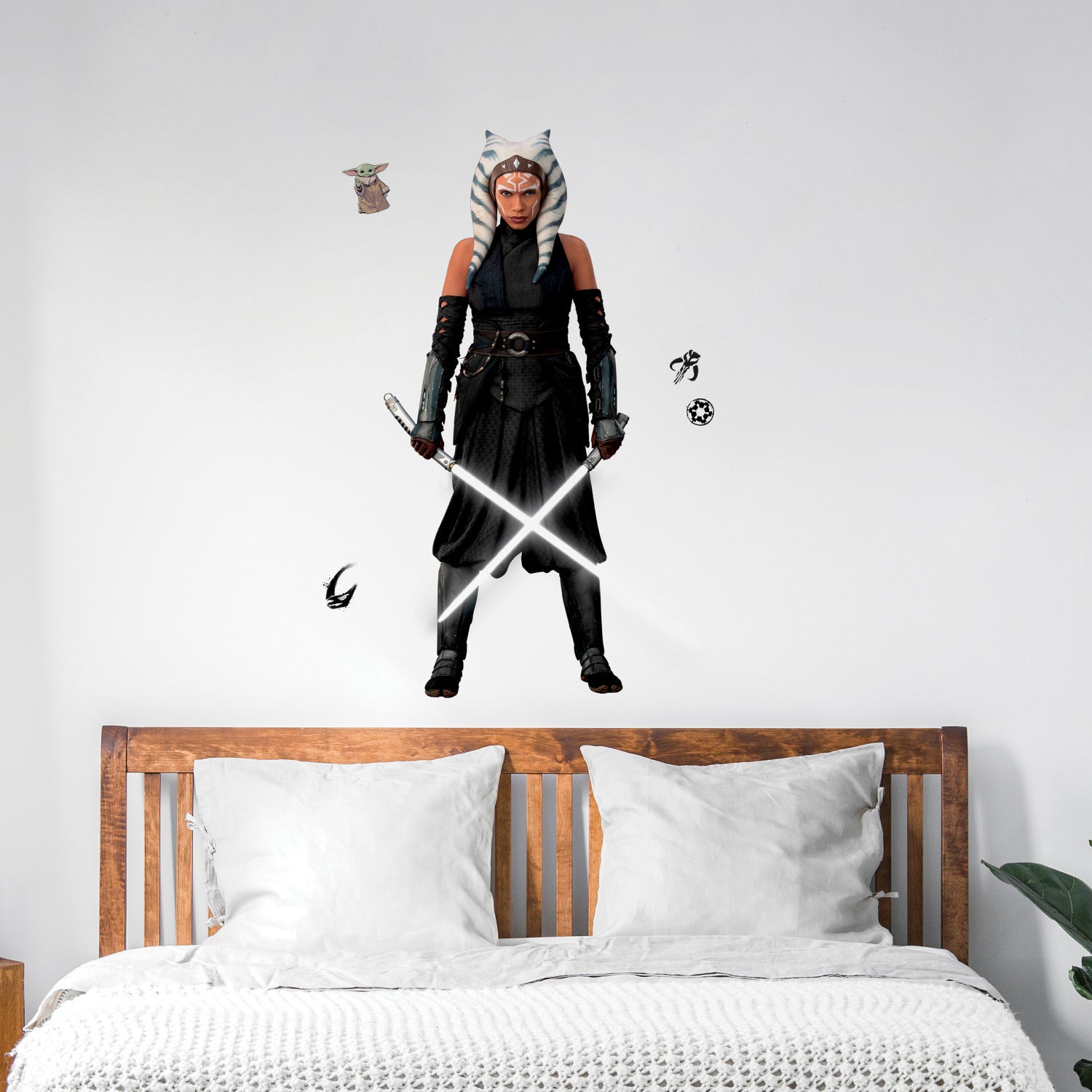A striking silhouette of Ahsoka Tano, clad in black armor with blue and white striped head-tails and facial markings, stands confidently holding two crossed white lightsabers—her arms wrapped in dark gauntlets—against a plain white background. The Star Wars Ahsoka Peel and Stick Giant Wall Decal by WallPops is an inspiring way to add bold character to any room.