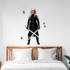 A striking silhouette of Ahsoka Tano, clad in black armor with blue and white striped head-tails and facial markings, stands confidently holding two crossed white lightsabers—her arms wrapped in dark gauntlets—against a plain white background. The Star Wars Ahsoka Peel and Stick Giant Wall Decal by WallPops is an inspiring way to add bold character to any room.