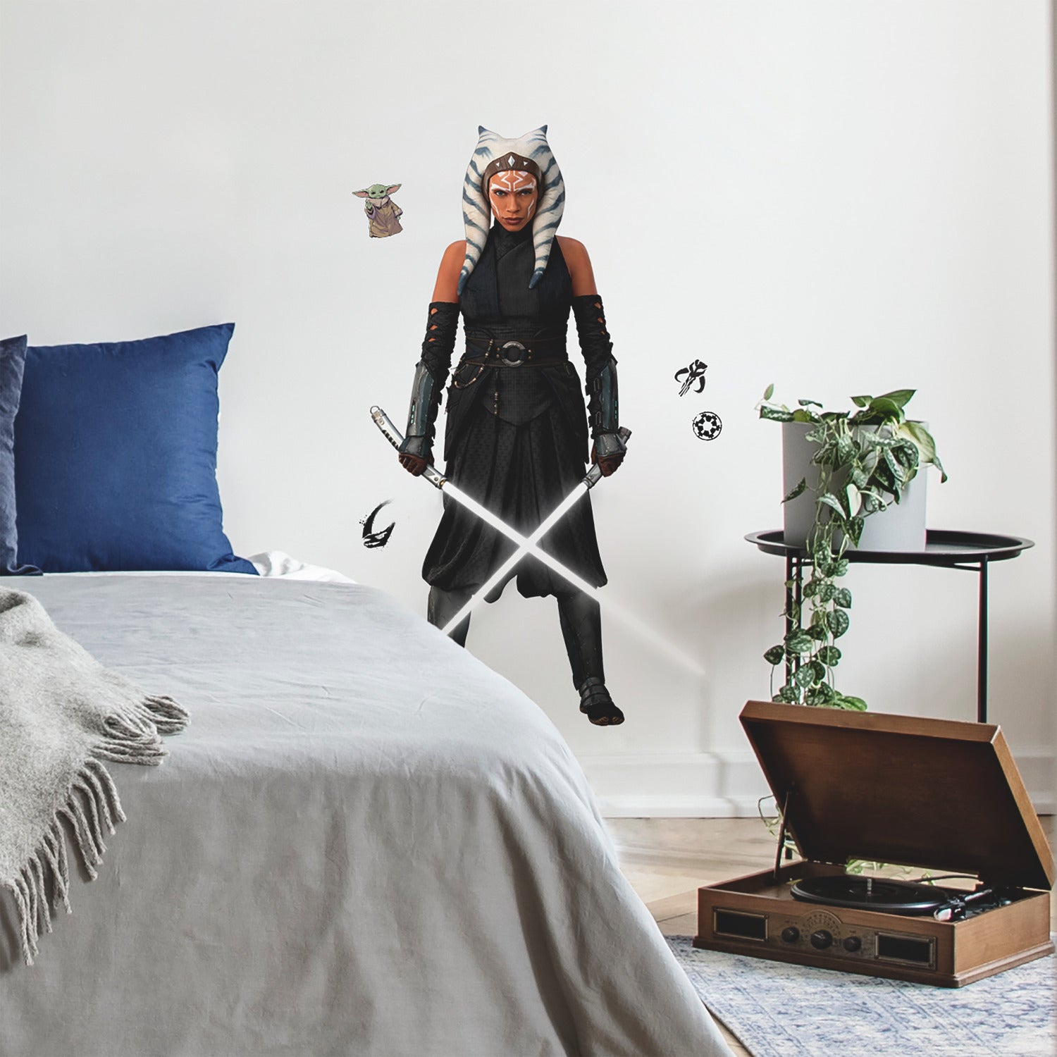 A striking silhouette of Ahsoka Tano, clad in black armor with blue and white striped head-tails and facial markings, stands confidently holding two crossed white lightsabers—her arms wrapped in dark gauntlets—against a plain white background. The Star Wars Ahsoka Peel and Stick Giant Wall Decal by WallPops is an inspiring way to add bold character to any room.