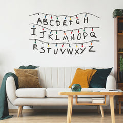 The WallPops Netflix Stranger Things Christmas Lights Peel And Stick Giant Wall Decals With Alphabet features a hand-drawn alphabet arranged in three rows, each row displayed beneath a line of colorful Christmas lights and accented with circled numbers 1–8 along the lines. This rustic design, measuring 27.43 69.6 cm tall by 46.04 1.16 m wide, boasts a light gray background reminiscent of distressed barn wood for added character.