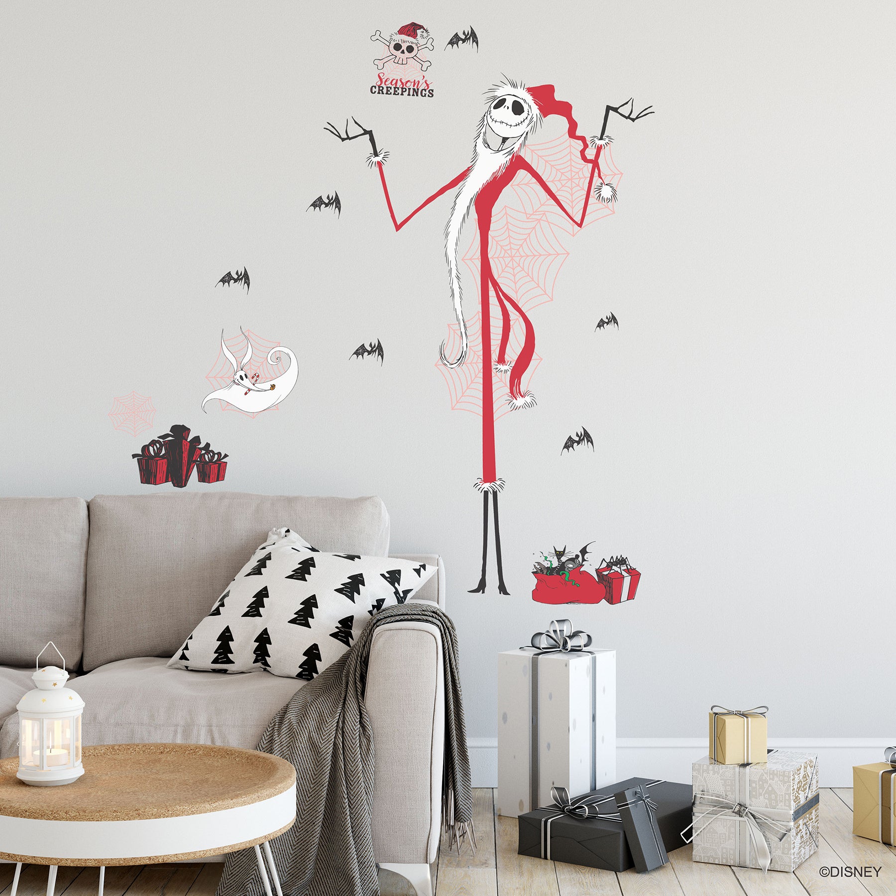 Giant Disney Tim Burtons The Nightmare Before Christmas Holiday Wall Decals by WallPops, featuring Jack Skellington, Zero, bats, spider webs, skulls, and presents, decorate the living room wall above a couch adorned with tree pillows. Rustic barn wood accents enhance the festive scene, while wrapped gifts, a lantern, and a blanket on the floor create a cozy blend of Halloween and Christmas décor.