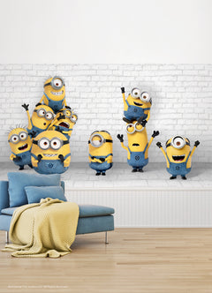 Eight Minions in blue overalls, with their signature yellow bodies and goggles, stand and stack joyfully against a white brick wall in the WallPops Minions Making Mischief Peel and Stick Mural. Some raise their arms in excitement, one perches playfully on top of another, and all display cheerful expressions.