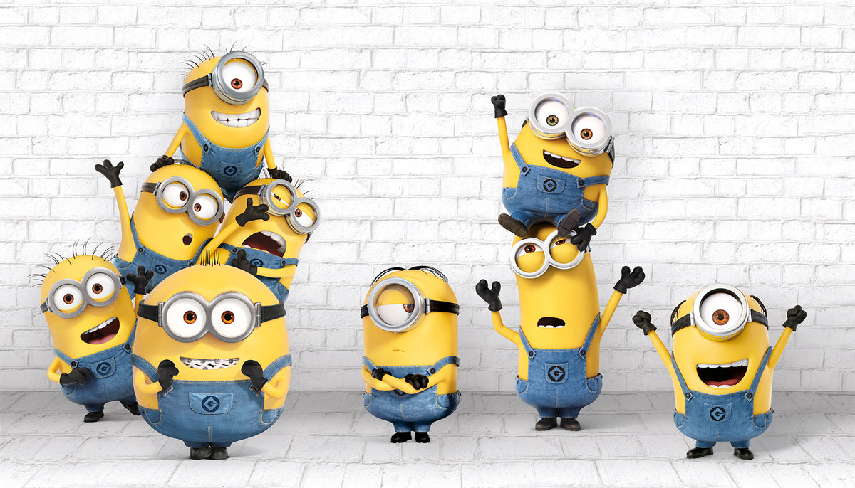 Eight Minions in blue overalls, with their signature yellow bodies and goggles, stand and stack joyfully against a white brick wall in the WallPops Minions Making Mischief Peel and Stick Mural. Some raise their arms in excitement, one perches playfully on top of another, and all display cheerful expressions.