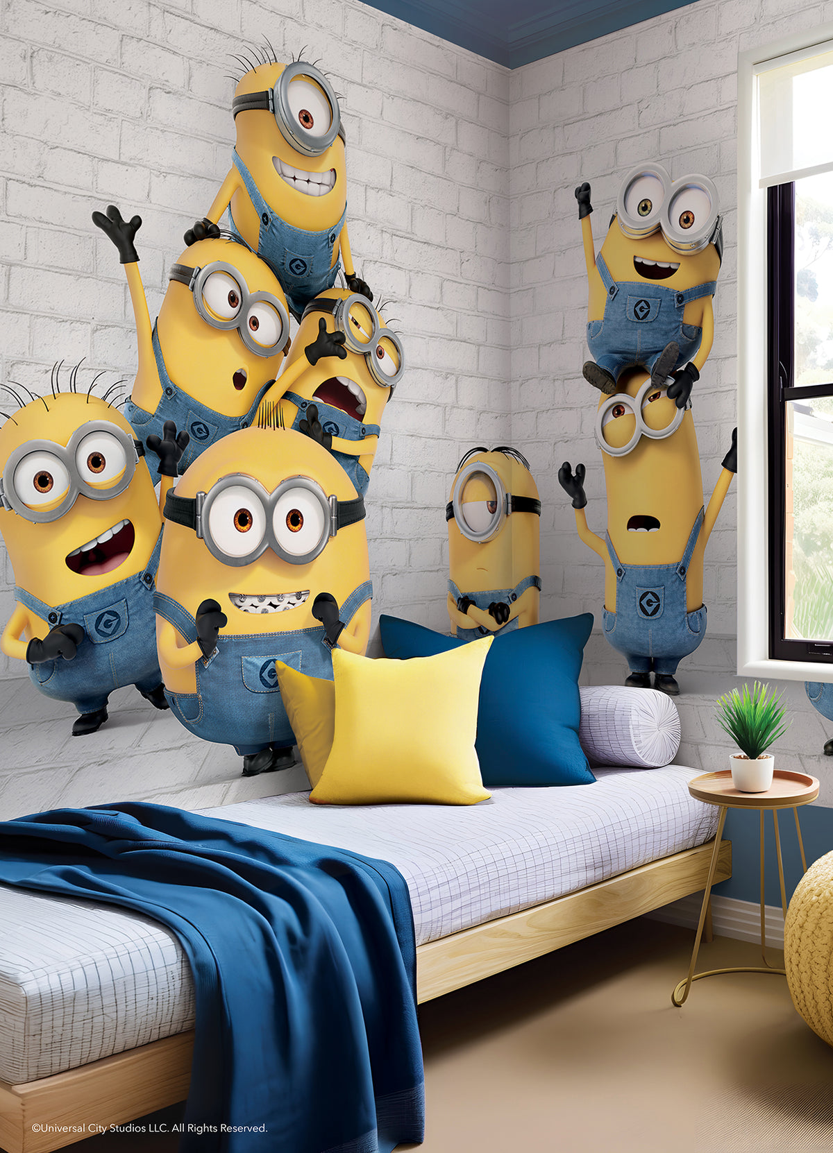 Eight Minions in blue overalls, with their signature yellow bodies and goggles, stand and stack joyfully against a white brick wall in the WallPops Minions Making Mischief Peel and Stick Mural. Some raise their arms in excitement, one perches playfully on top of another, and all display cheerful expressions.