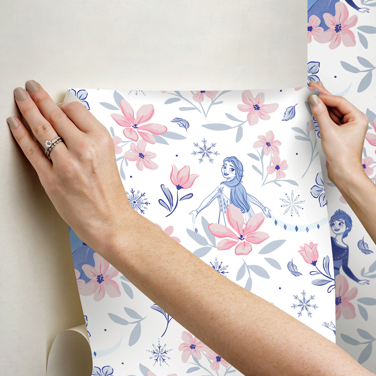 Hands with a silver ring smooth Frozen II Frozen 2 Anna and Elsa Floral Peel and Stick Wallpaper by WallPops onto a cream wall, aligning its repeating pink floral pattern and cartoon girls in blue dresses.