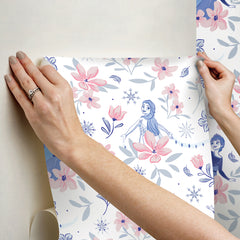 Hands with a silver ring smooth Frozen II Frozen 2 Anna and Elsa Floral Peel and Stick Wallpaper by WallPops onto a cream wall, aligning its repeating pink floral pattern and cartoon girls in blue dresses.