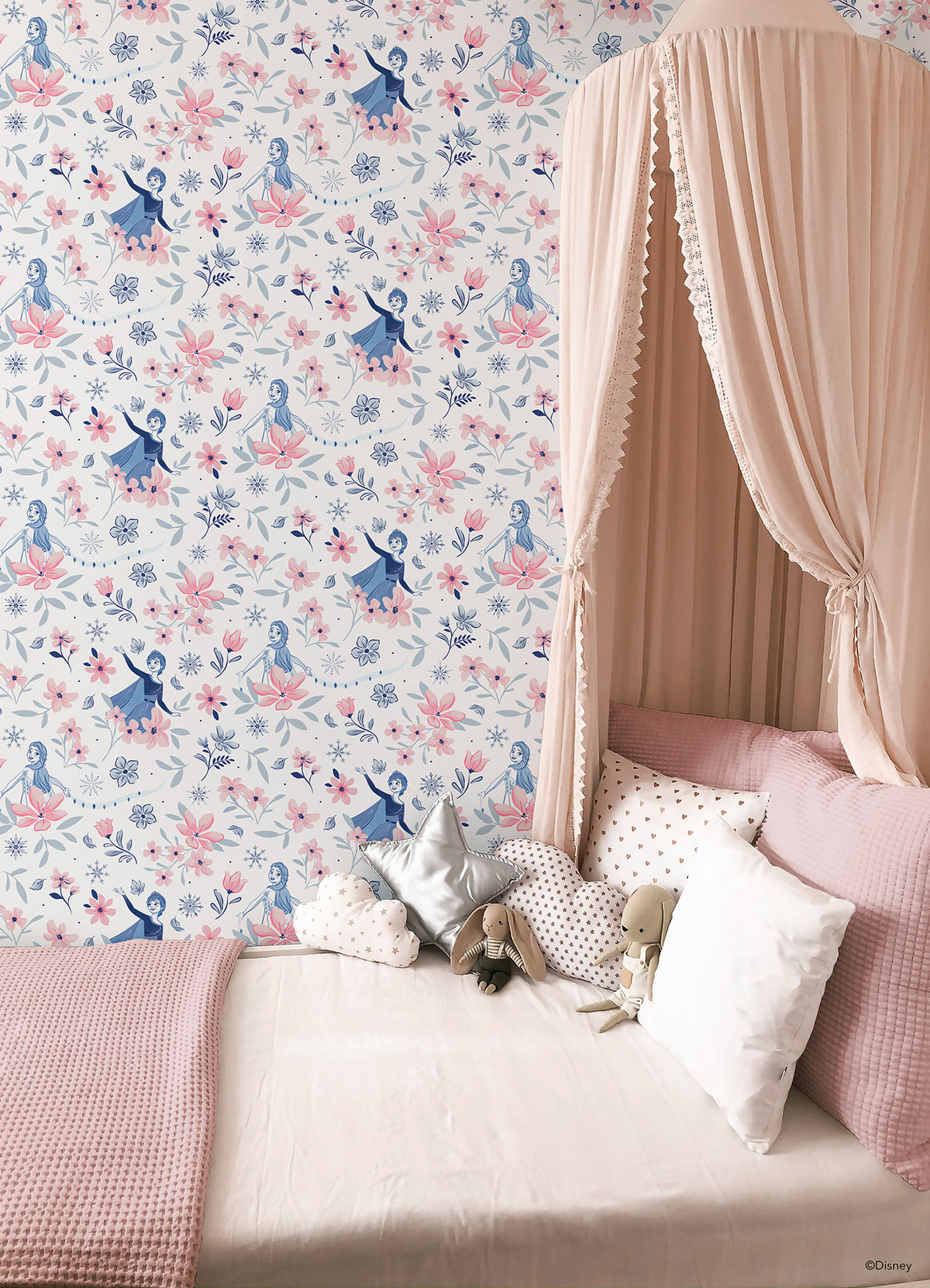 Hands with a silver ring smooth Frozen II Frozen 2 Anna and Elsa Floral Peel and Stick Wallpaper by WallPops onto a cream wall, aligning its repeating pink floral pattern and cartoon girls in blue dresses.