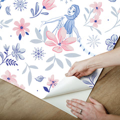 Hands with a silver ring smooth Frozen II Frozen 2 Anna and Elsa Floral Peel and Stick Wallpaper by WallPops onto a cream wall, aligning its repeating pink floral pattern and cartoon girls in blue dresses.