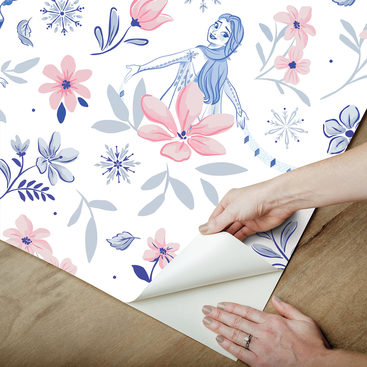 Hands with a silver ring smooth Frozen II Frozen 2 Anna and Elsa Floral Peel and Stick Wallpaper by WallPops onto a cream wall, aligning its repeating pink floral pattern and cartoon girls in blue dresses.