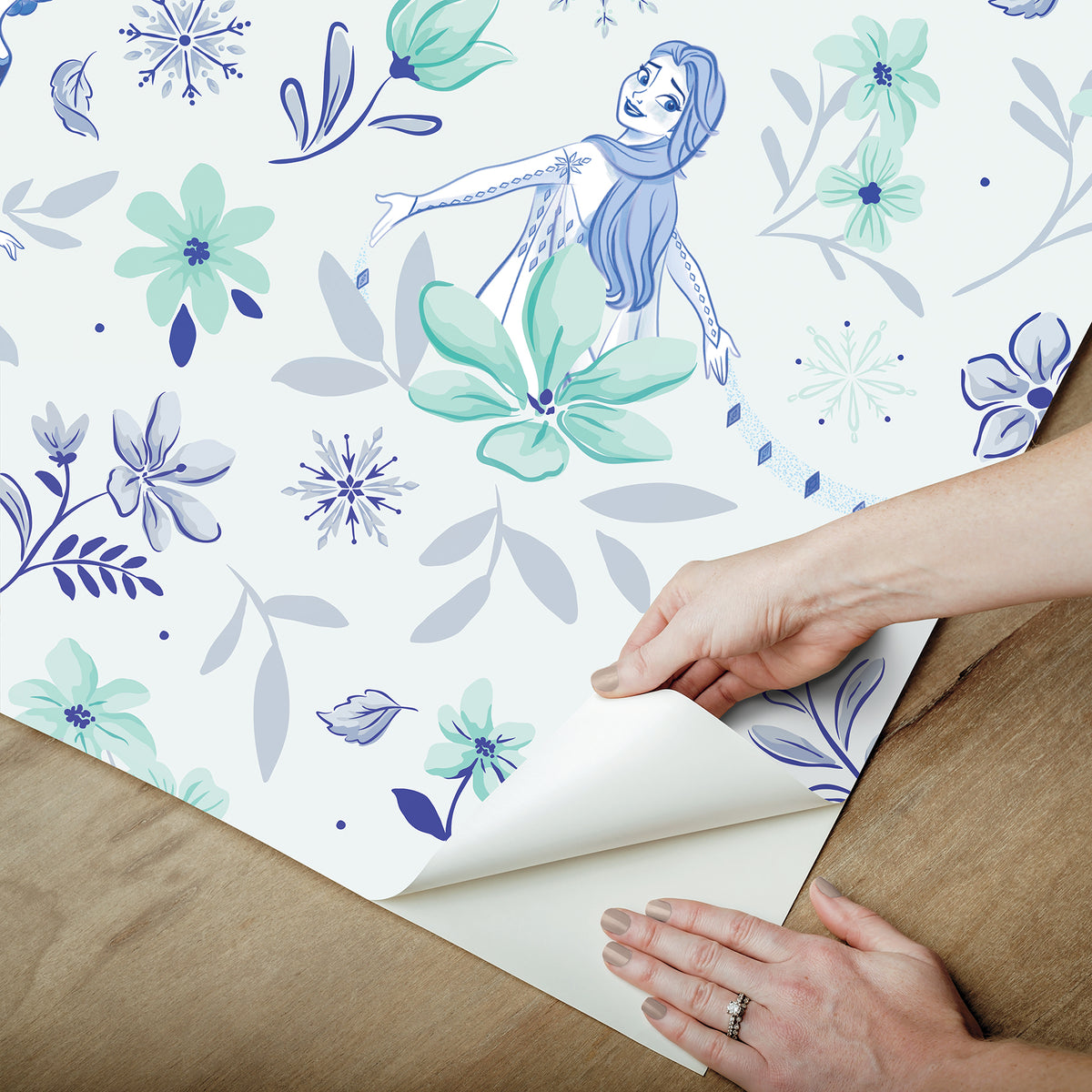Hands with a silver ring smooth Frozen II Frozen 2 Anna and Elsa Floral Peel and Stick Wallpaper by WallPops onto a cream wall, aligning its repeating pink floral pattern and cartoon girls in blue dresses.