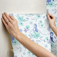 Hands with a silver ring smooth Frozen II Frozen 2 Anna and Elsa Floral Peel and Stick Wallpaper by WallPops onto a cream wall, aligning its repeating pink floral pattern and cartoon girls in blue dresses.