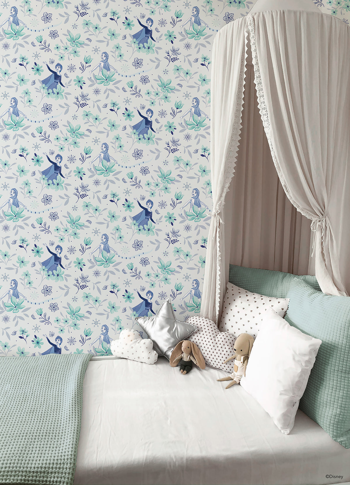 Hands with a silver ring smooth Frozen II Frozen 2 Anna and Elsa Floral Peel and Stick Wallpaper by WallPops onto a cream wall, aligning its repeating pink floral pattern and cartoon girls in blue dresses.