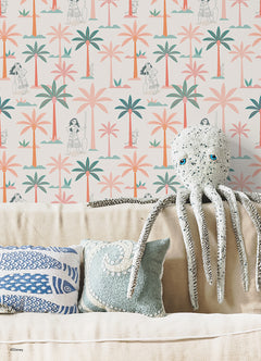 Two twin beds with white bedding flank a white nightstand, which holds a lamp, plant, and books. The WallPops Disney Moana Coconut Tree Peel and Stick Wallpaper showcases green and pink palm trees mixed with small black-and-white illustrations of women, bringing an artistic flair to the space. The overall look is fresh, playful, and tropical.