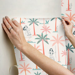 Two twin beds with white bedding flank a white nightstand, which holds a lamp, plant, and books. The WallPops Disney Moana Coconut Tree Peel and Stick Wallpaper showcases green and pink palm trees mixed with small black-and-white illustrations of women, bringing an artistic flair to the space. The overall look is fresh, playful, and tropical.
