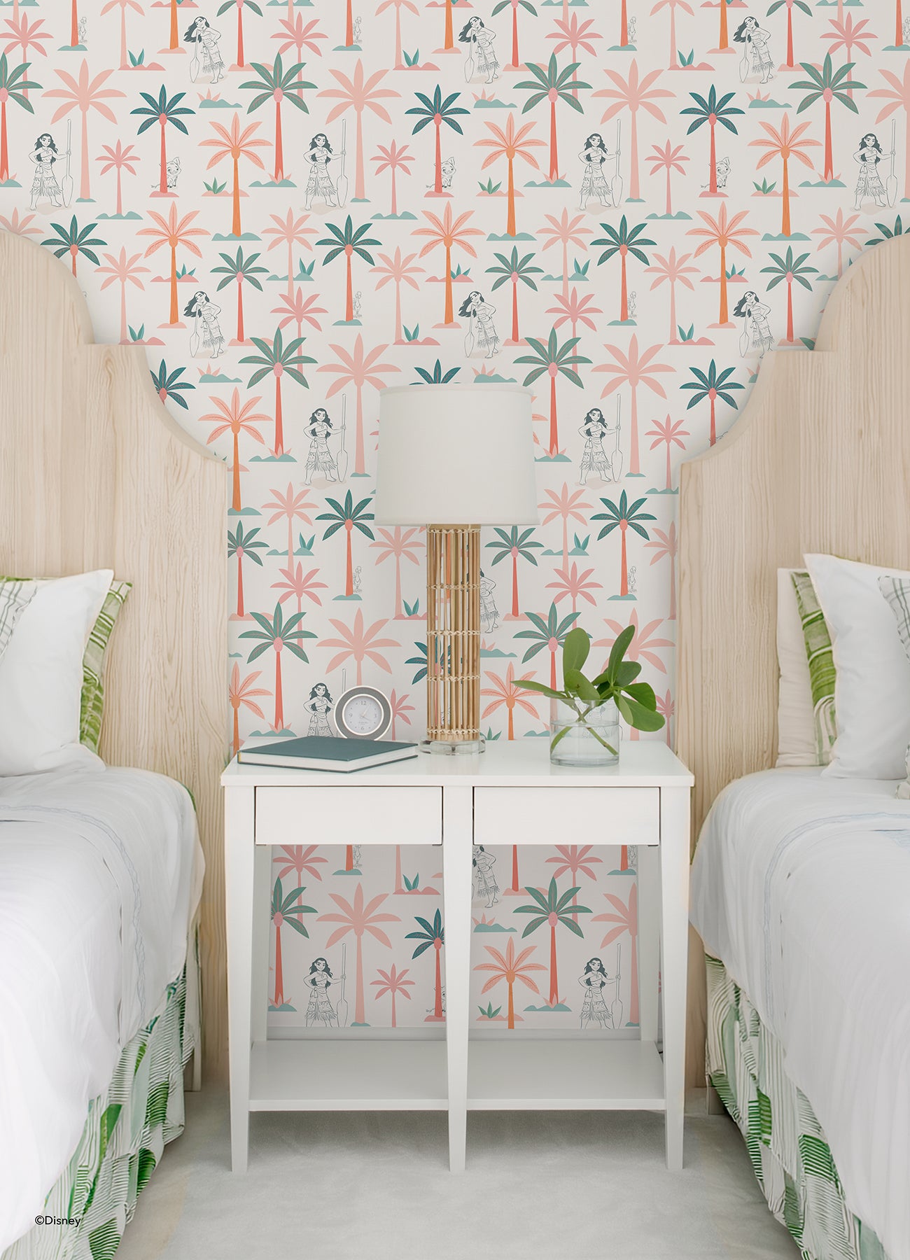 Two twin beds with white bedding flank a white nightstand, which holds a lamp, plant, and books. The WallPops Disney Moana Coconut Tree Peel and Stick Wallpaper showcases green and pink palm trees mixed with small black-and-white illustrations of women, bringing an artistic flair to the space. The overall look is fresh, playful, and tropical.