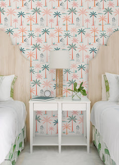 Two twin beds with white bedding flank a white nightstand, which holds a lamp, plant, and books. The WallPops Disney Moana Coconut Tree Peel and Stick Wallpaper showcases green and pink palm trees mixed with small black-and-white illustrations of women, bringing an artistic flair to the space. The overall look is fresh, playful, and tropical.