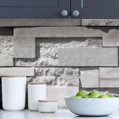 A grayscale photo features a wall covered with WallPops Mineral Siding Adhesive Film, designed to mimic staggered rectangular bricks. The film displays a variety of surfaces: some areas are smooth, while others have rough textures or subtle floral engravings, creating striking contrasts in patterns and finishes. Soft lighting casts gentle shadows, highlighting the realistic relief of the Mineral Siding design.