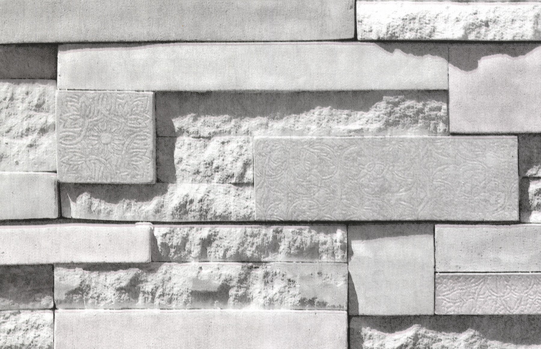 A grayscale photo features a wall covered with WallPops Mineral Siding Adhesive Film, designed to mimic staggered rectangular bricks. The film displays a variety of surfaces: some areas are smooth, while others have rough textures or subtle floral engravings, creating striking contrasts in patterns and finishes. Soft lighting casts gentle shadows, highlighting the realistic relief of the Mineral Siding design.