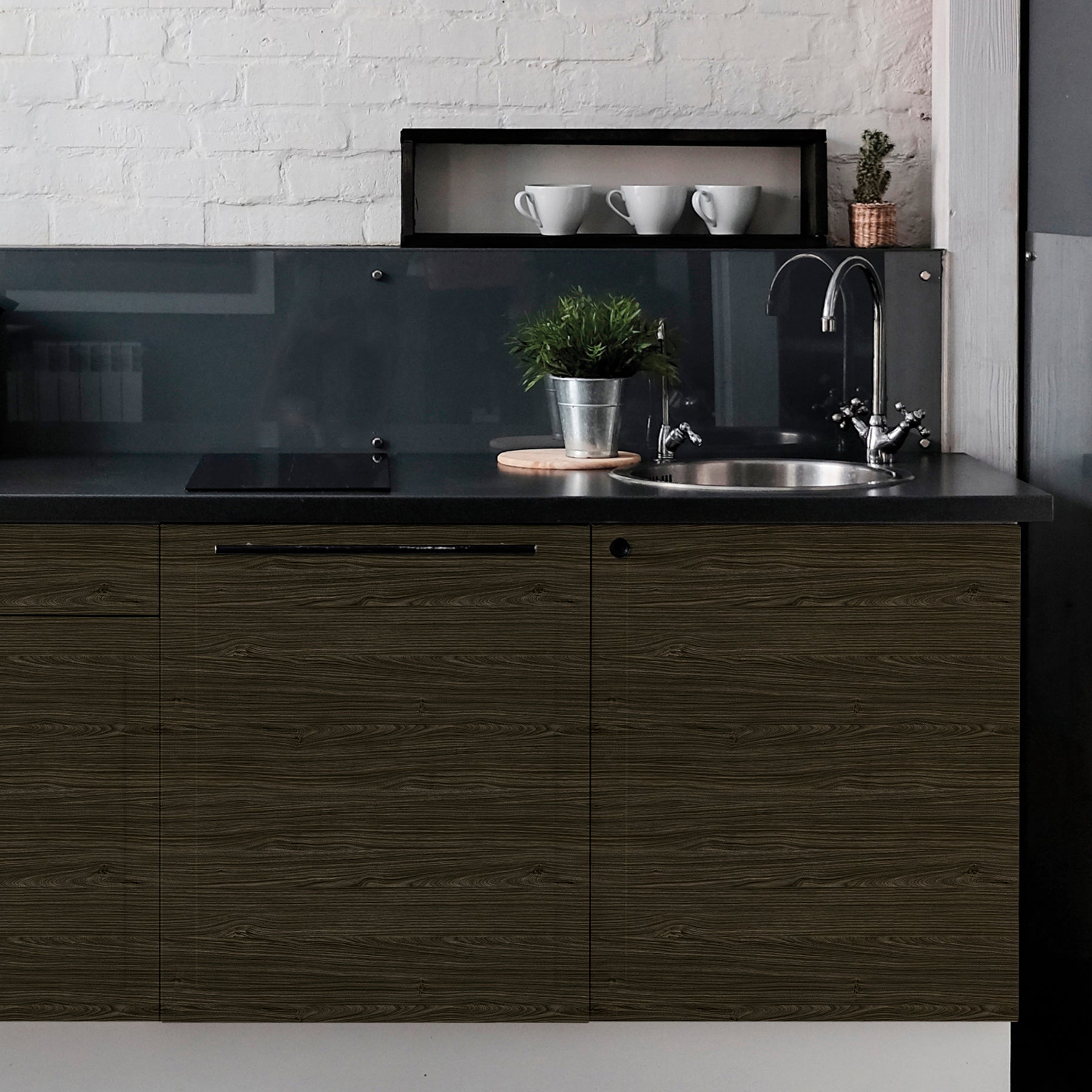 A modern kitchen featuring WallPops Dark Ash Adhesive Film on the cabinets, a round metal sink, a potted plant, a small cactus, three white cups, and a glass backsplash. The wall behind is finished in white brick, complemented by a black countertop and soft lighting for a cozy atmosphere.