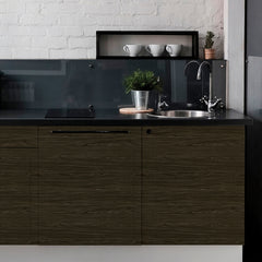 A modern kitchen featuring WallPops Dark Ash Adhesive Film on the cabinets, a round metal sink, a potted plant, a small cactus, three white cups, and a glass backsplash. The wall behind is finished in white brick, complemented by a black countertop and soft lighting for a cozy atmosphere.