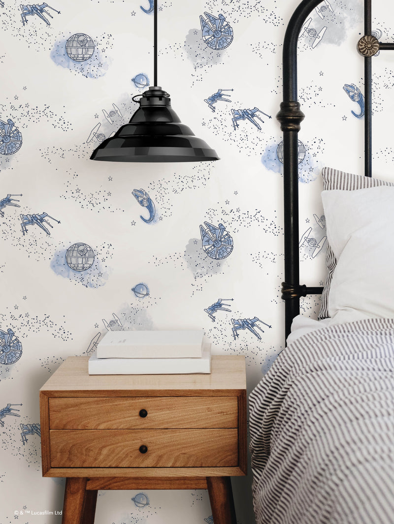 A wooden nightstand with books sits beside a black metal bed with striped bedding. A black pendant lamp hangs above. The wallpaper features blue Star Wars ships, droids, and stars on a white background.