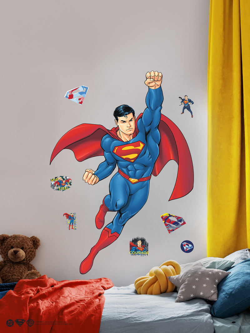 A colorful wall decal of Superman flying with one fist raised is above a bed with yellow curtains, a star-patterned pillow, a knit yellow cushion, a teddy bear, and smaller superhero stickers on the wall. The room has a playful, child-friendly atmosphere.