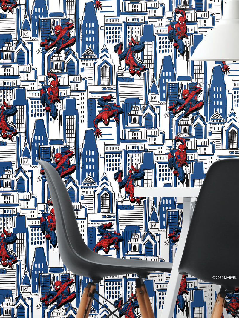 A modern dining area with white and black chairs sits before wallpaper featuring multiple Spider-Man illustrations swinging among blue and white city skyscrapers. The dynamic poses of Spider-Man contrast with the geometric urban background.